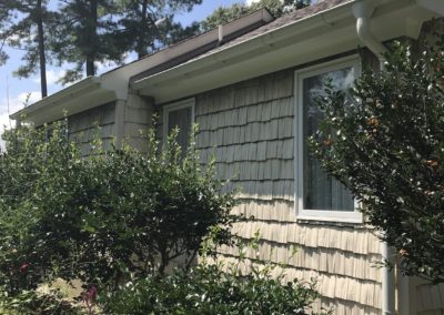 Two gutter sections. Staggered installation. Light beige shake siding and lots of green bushes in the landscaping.