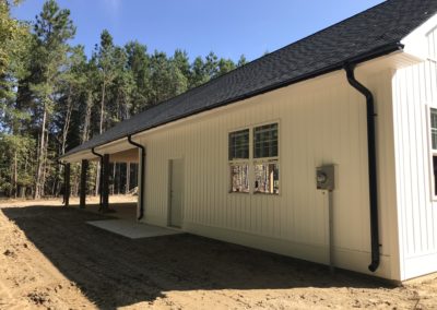 Backside of workshop building. White siding. Black gutters and downspouts. Blue sky and shadow on the ground from the building.